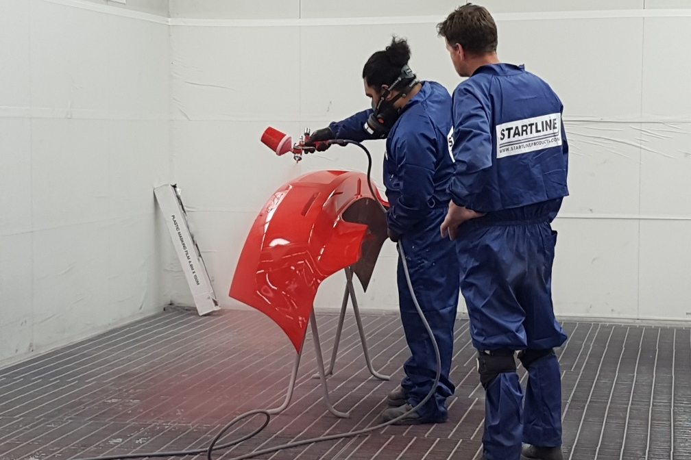 Spray painter applying colour to a red bumper in a spray booth